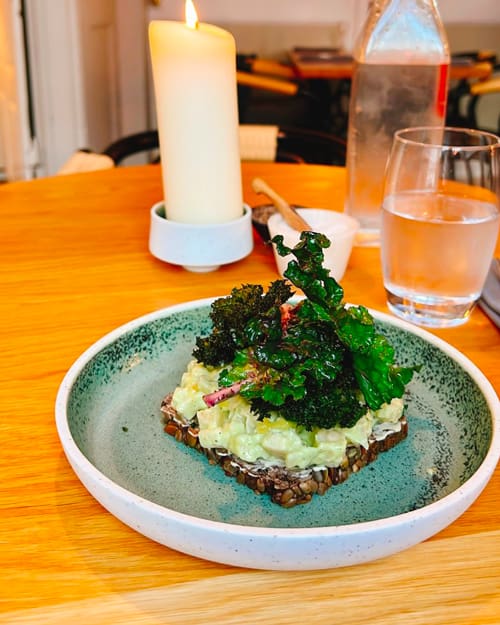 Seeded toast with avocado spread and kale garnish on a ceramic plate, candlelit table in background.