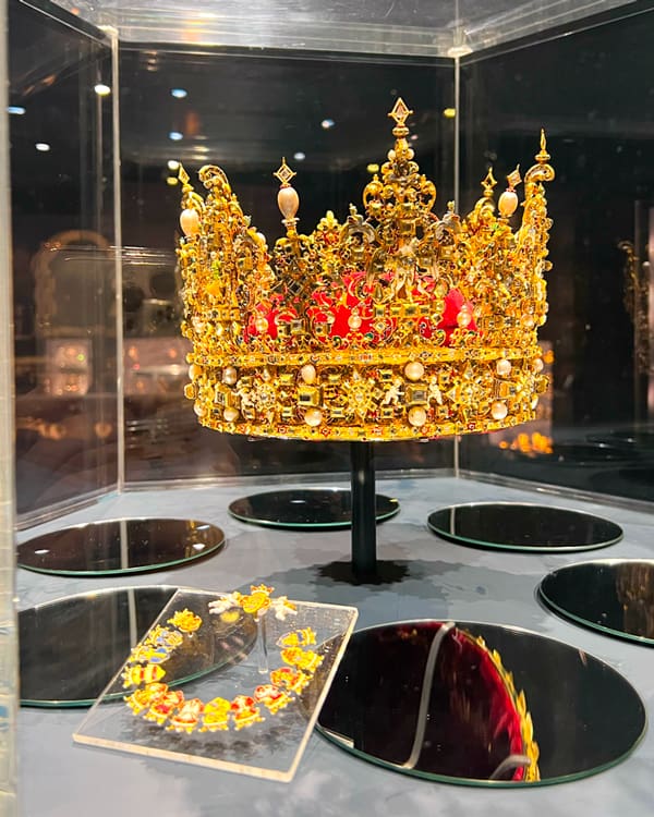Ornate gold crown encrusted with pearls and red gems on a black display stand inside a glass case.