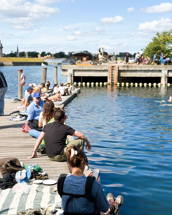 People sit along a wooden dock by a blue lake on a sunny day, some fishing or watching the water.