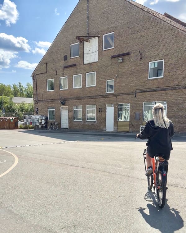 Rear view of a person cycling on a sunny street past a brick multi‑story building with white doors and windows.