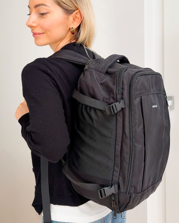 Side view of a woman wearing a black backpack with multiple compartments and adjustable straps, standing indoors.