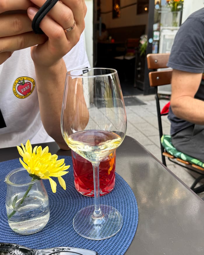 Wine glass on a blue placemat at an outdoor cafe, with a small vase of yellow flower and a person resting their hands nearby.