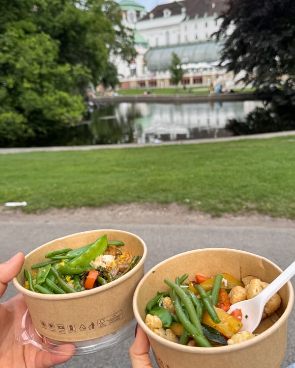 Two paper bowls of veggie stir-fry held in a park with a pond and historic buildings in the background.