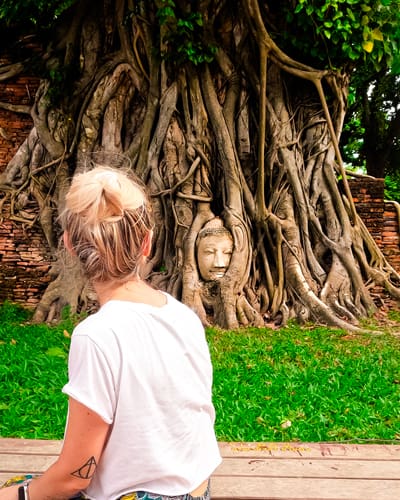 Person with a bun and white T-shirt stands before a massive tree with exposed roots; a Buddha head is carved into the trunk.