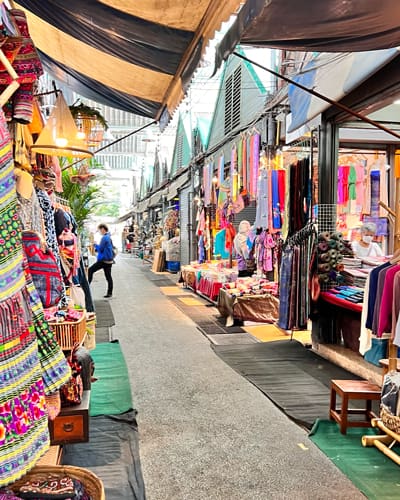 Outdoor market street with colorful fabric stalls and clothing racks; shoppers browse along a narrow walkway between vendors.