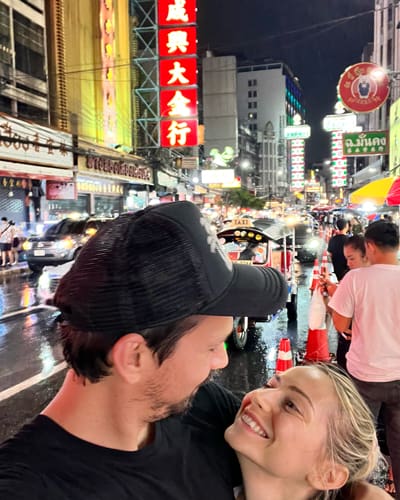Couple smiling as they pose for a selfie on a crowded neon-lit street at night in a busy Asian city, with bright signs and pedestrians in the background.