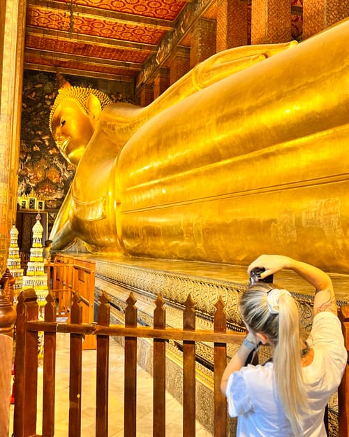 Large golden reclining Buddha statue inside an ornate temple; a visitor photographs it with a phone.