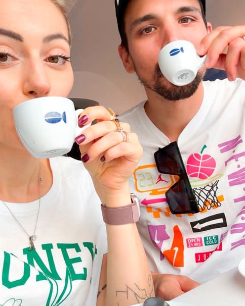 Two friends share a close-up selfie while sipping espresso from white cups with a blue logo; woman on left with pink nails and a watch, man on right wearing a graphic white shirt and sunglasses hanging from his neckline.
