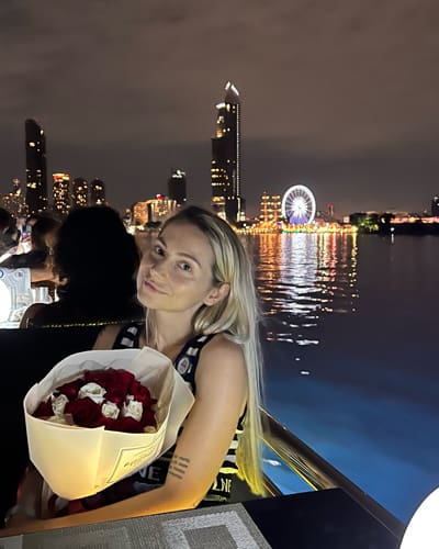 Woman on a night boat ride holding a bouquet of red roses, with a lit city skyline and ferris wheel behind the scene.