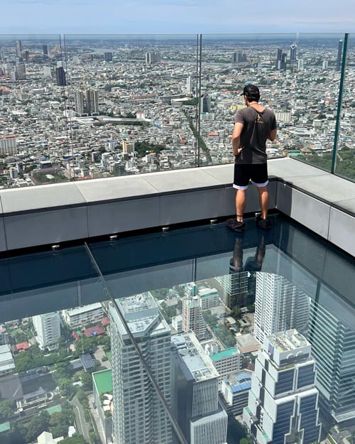 Man in a gray shirt and black shorts stands at the edge of a glass-floored observation deck, overlooking a city.