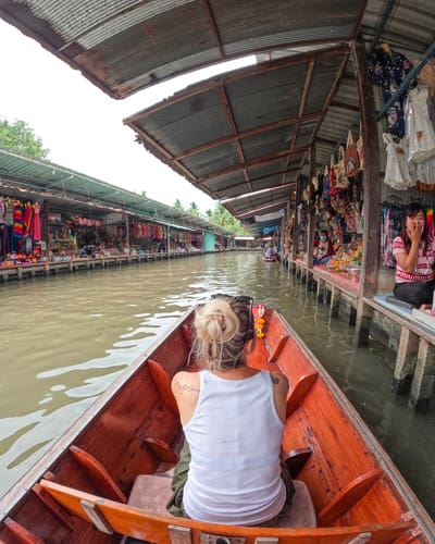 Person with light hair in a white sleeveless shirt sits in a wooden boat, gliding along a canal flanked by market stalls selling clothes.