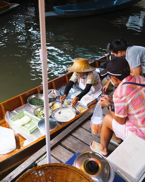 Vendor in a straw hat cooks fresh food on a wooden boat market, customers nearby selecting ingredients by the water's edge