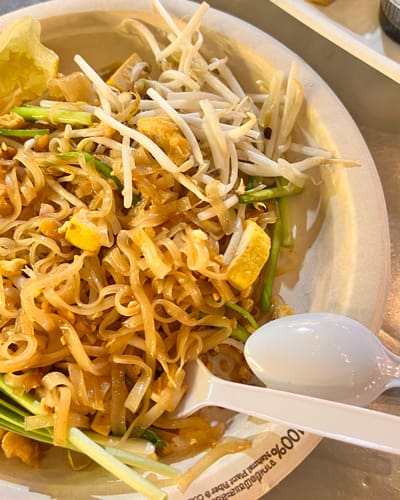 Stir-fried noodles with bean sprouts, tofu, and green onions on a plate with a white spoon nearby.