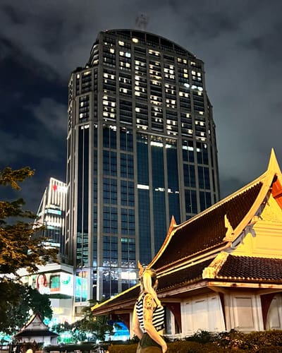 Night cityscape showing a tall glass skyscraper beside a traditional curved-roof building, with a person in a striped shirt in the foreground.