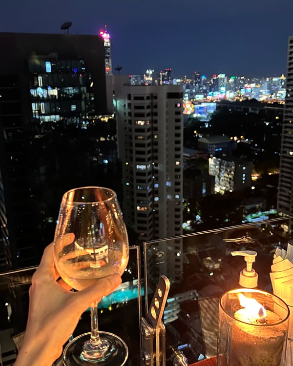 Nighttime city skyline viewed from a balcony; a hand holds a wine glass in the foreground, with a lit candle and pump bottle on the railing.