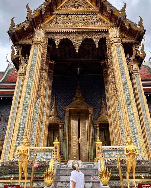 Ornate golden temple entrance with intricate blue tilework, flanked by gold statues, as a person in a white shirt faces the doorway.