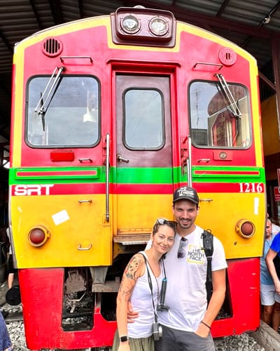 Couple posing and smiling in front of a bright red and yellow locomotive with a green stripe inside a rail yard.
