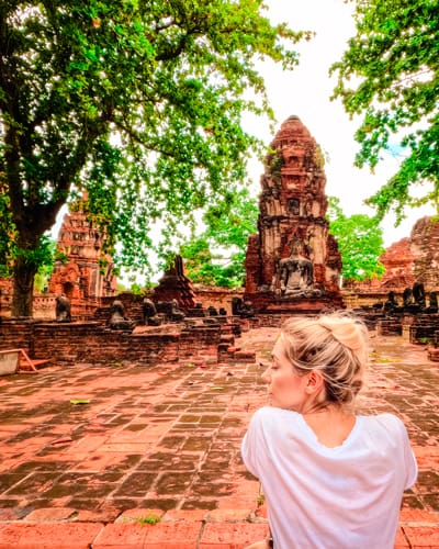 Woman with blonde hair in a white shirt stands with her back to the camera, facing ancient brick temple ruins amid green trees.