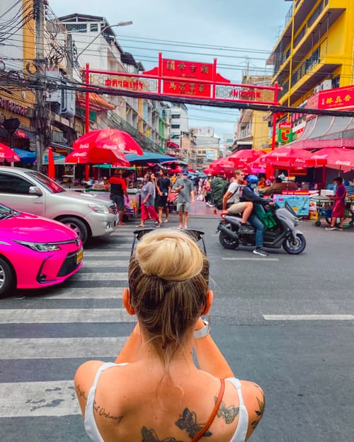Back view of a woman with a bun standing at a crosswalk, facing a busy street market with red umbrellas, scooters, and colorful buildings.