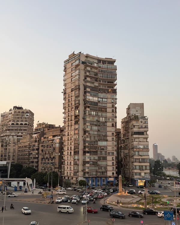 Cluster of tall, cylindrical apartment towers lining a busy city intersection at sunset with cars on the road and a pale sky behind them