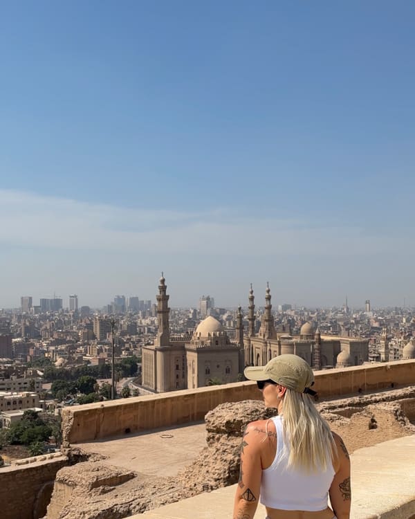 Woman with blonde hair in a white tank top and cap stands on a rooftop, overlooking Cairo's skyline with mosques and domes in the distance, clear blue sky above.