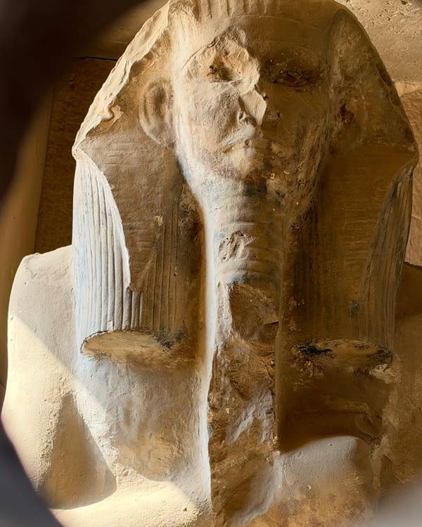 Stone statue of an ancient Egyptian pharaoh with a nemes headdress and weathered face, carved from light-colored rock, in a dusty setting.