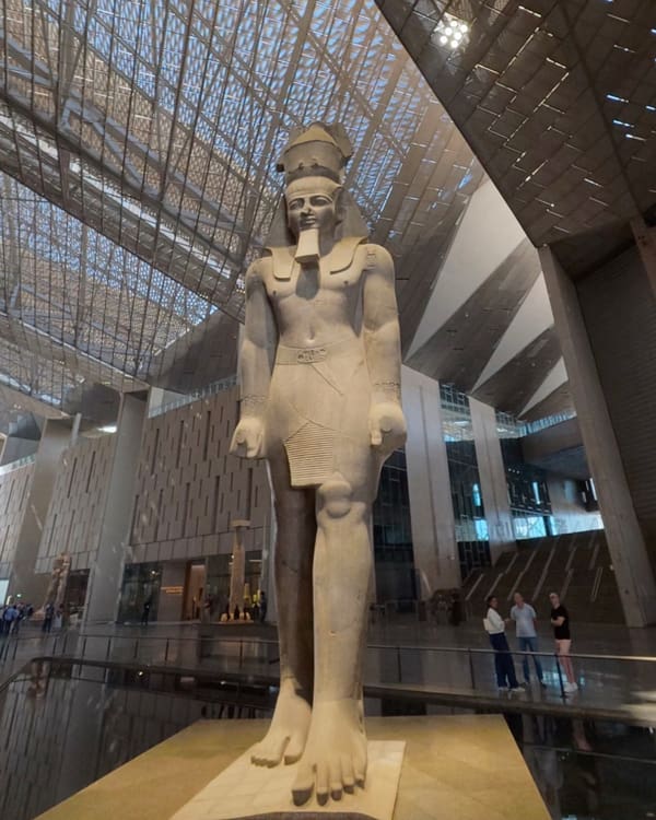 Ancient Egyptian statue of a pharaoh standing with arms at sides, wearing a tall headdress, on a pedestal inside a modern museum hall with glass ceiling.