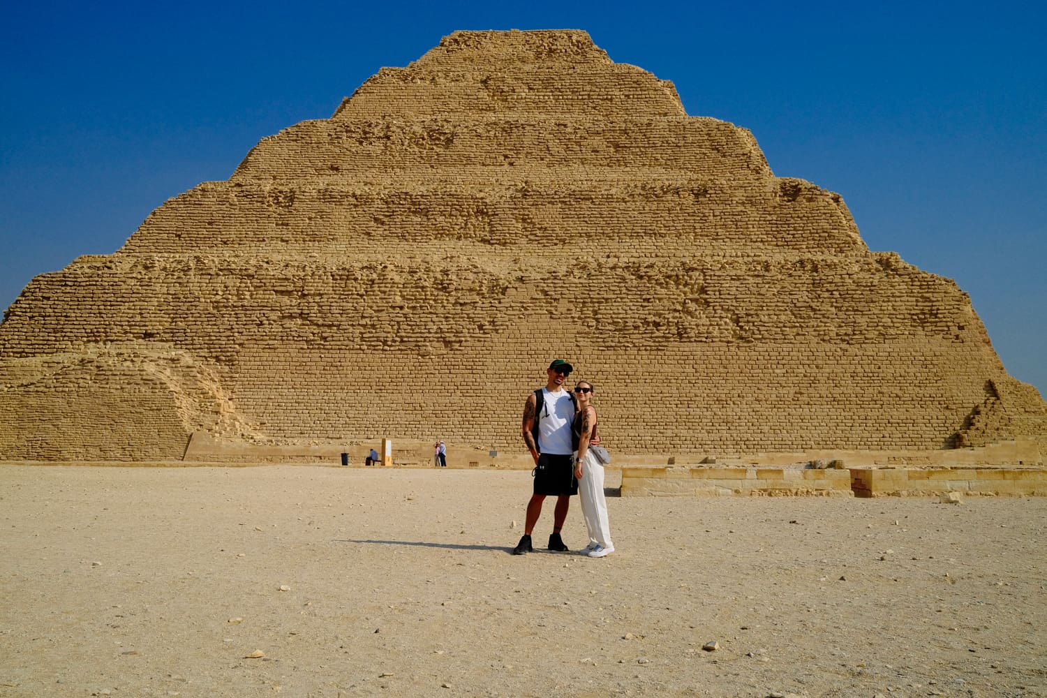 Two tourists stand together in front of a massive ancient pyramid in a desert under a clear blue sky, posing for a photo.
