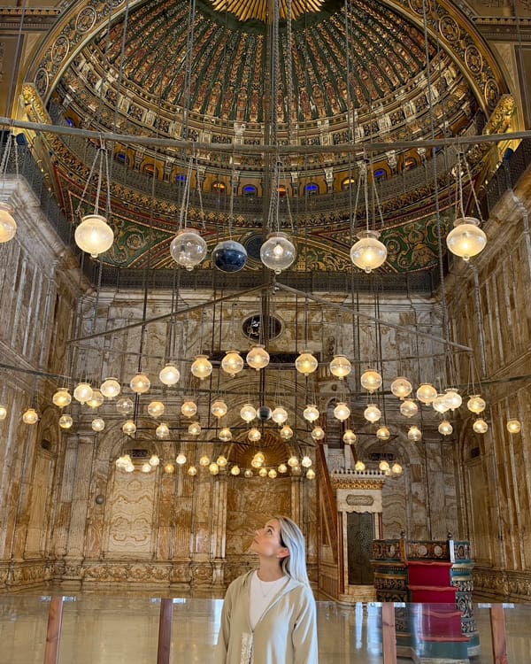Woman in light coat looks up at many hanging glass globes inside a richly decorated domed hall with marble walls and ornate ceiling.