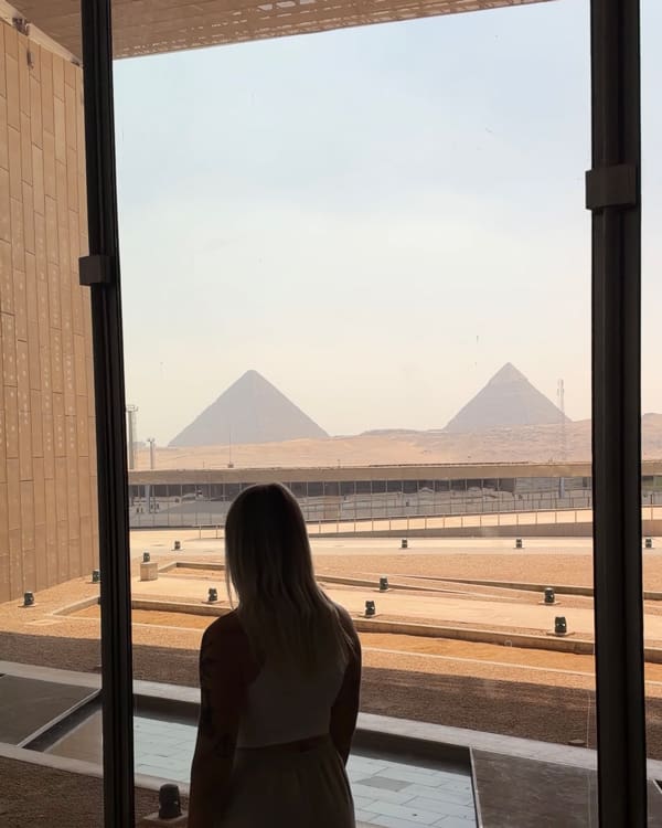 Silhouette of a woman in a white dress standing inside a glass-walled building, looking toward two pyramids on a desert plain.