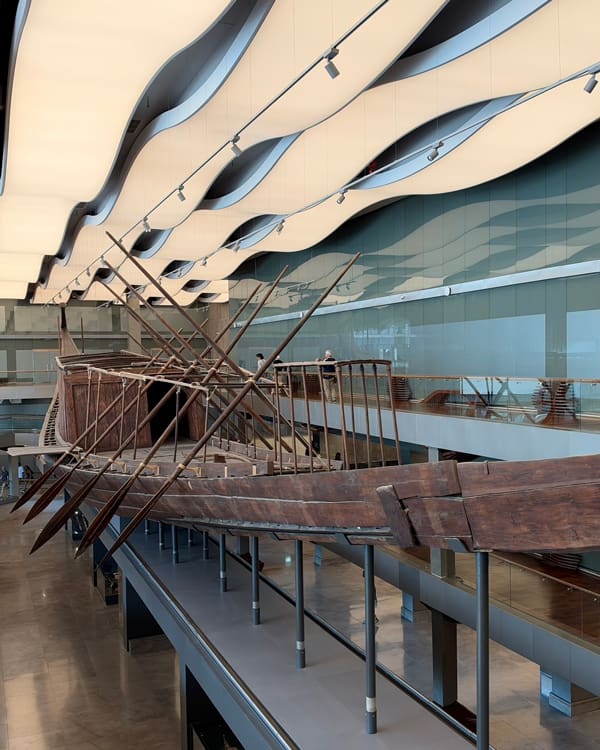 Large wooden ship sculpture displayed indoors under a wavy white ceiling in a museum atrium.