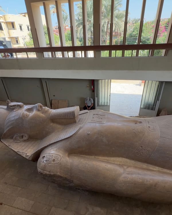 Large reclining ancient Egyptian statue of a pharaoh in a workshop, with a worker in the background and open doors behind it.